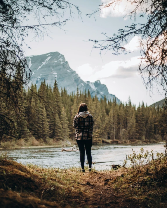 a person standing in front of a body of water, rocky mountains and a river, enjoying a stroll in the forest, looking sad, banff national park