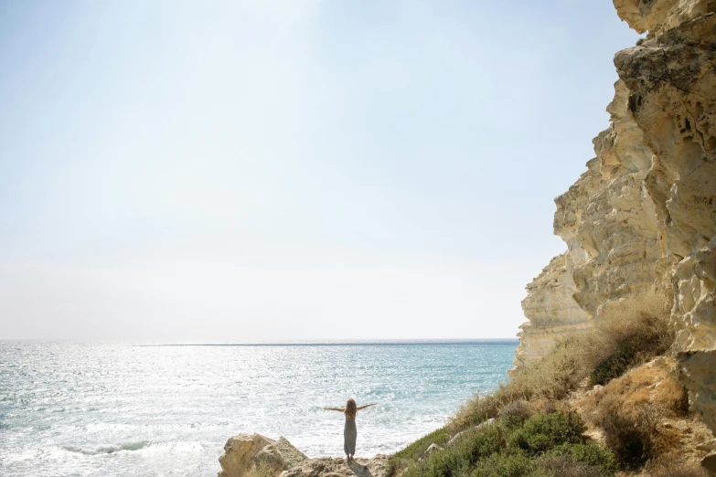 a man standing on top of a cliff next to the ocean, by Will Ellis, pexels contest winner, minimalism, cyprus, margot robbie on the beach, spreading her wings, an olive skinned