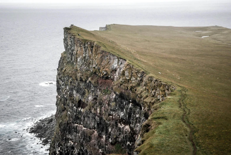 a sheep standing on top of a cliff next to the ocean, by Þórarinn B. Þorláksson, pexels contest winner, les nabis, tiny people walking below, gigapixel photo, grey, brown
