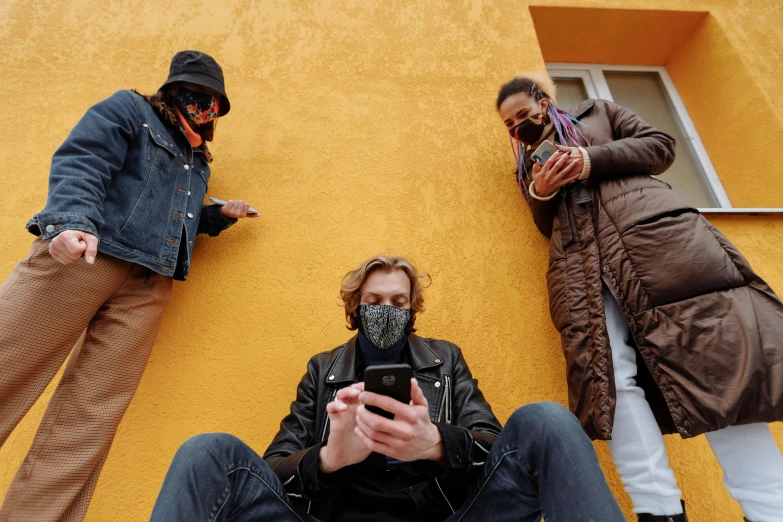 three people standing next to each other in front of a yellow building, trending on pexels, street art, white man with black fabric mask, cell phones, programming, brown