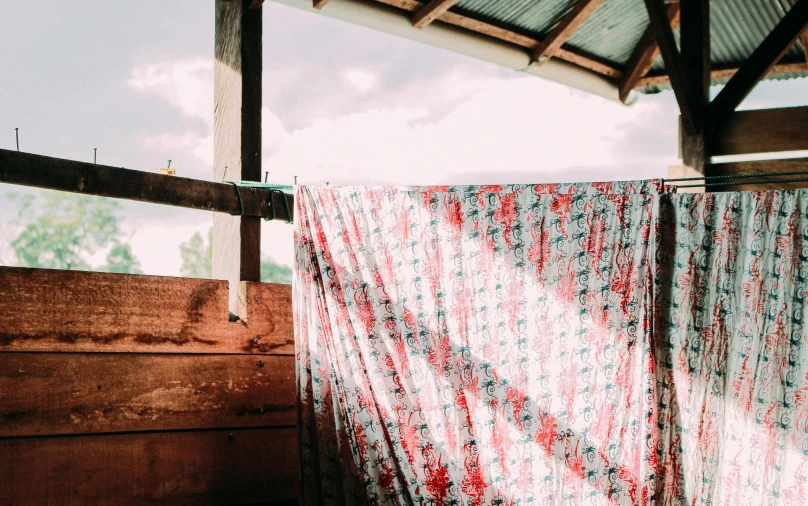 a bed sitting under a metal roof next to a window, a silk screen, by Pamela Ascherson, unsplash, sarong, on a farm, red fabric, blocking the sun