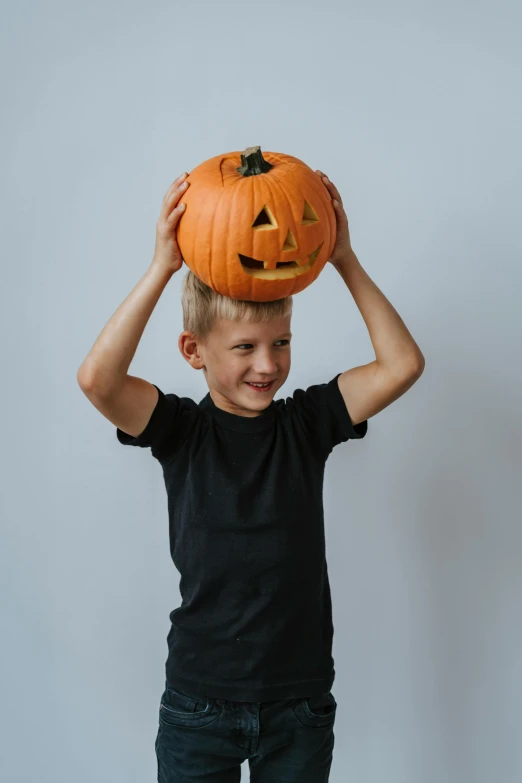 a young boy holding a pumpkin on his head, pexels contest winner, on a gray background, hip-length, dark. no text, scaring