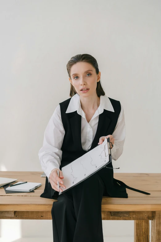 a woman sitting at a table holding a piece of paper, wearing a vest and a tie, white and black clothing, 2019 trending photo, gif