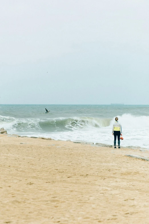 a man standing on top of a sandy beach next to the ocean, surfing, li zixin, omaha beach, rough waters