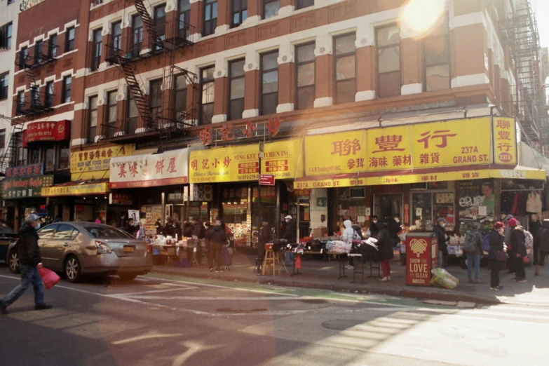 a group of people walking down a street next to tall buildings, chinatown, storefront, sunny light, street corner