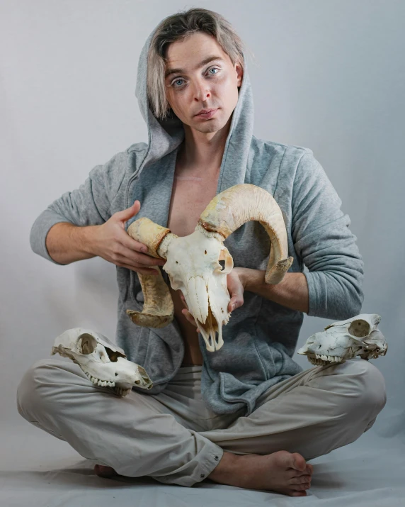 a man sitting on the ground holding a goat skull, by Jessie Alexandra Dick, unsplash, renaissance, silvery skinned male elf, full view of face and body, on display, non binary model