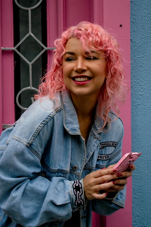 a woman with pink hair standing in front of a pink door, trending on pexels, happening, denim jacket, curly blonde hair | d & d, she is holding a smartphone, charli bowater and artgeem
