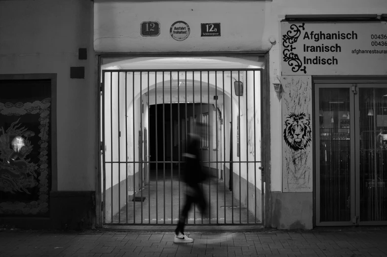 a black and white photo of a person walking in front of a building, by Kristian Zahrtmann, pexels contest winner, graffiti, gate, people running, late night, prisoner