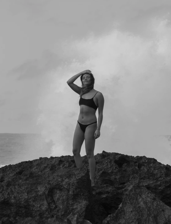 a woman standing on top of a rock next to the ocean, a black and white photo, by Robbie Trevino, made of lava, is wearing a swimsuit, josh grover, profile image