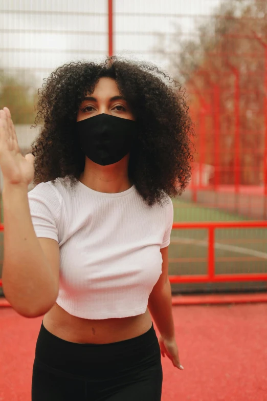 a woman standing on a soccer field wearing a face mask, pexels contest winner, long afro hair, wearing a cropped black tank top, hands shielding face, she is wearing streetwear