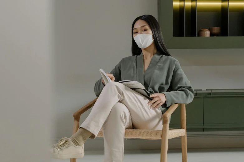 a woman sitting in a chair reading a book, inspired by Fei Danxu, facemask, beige, clean aesthetic, clean medical environment