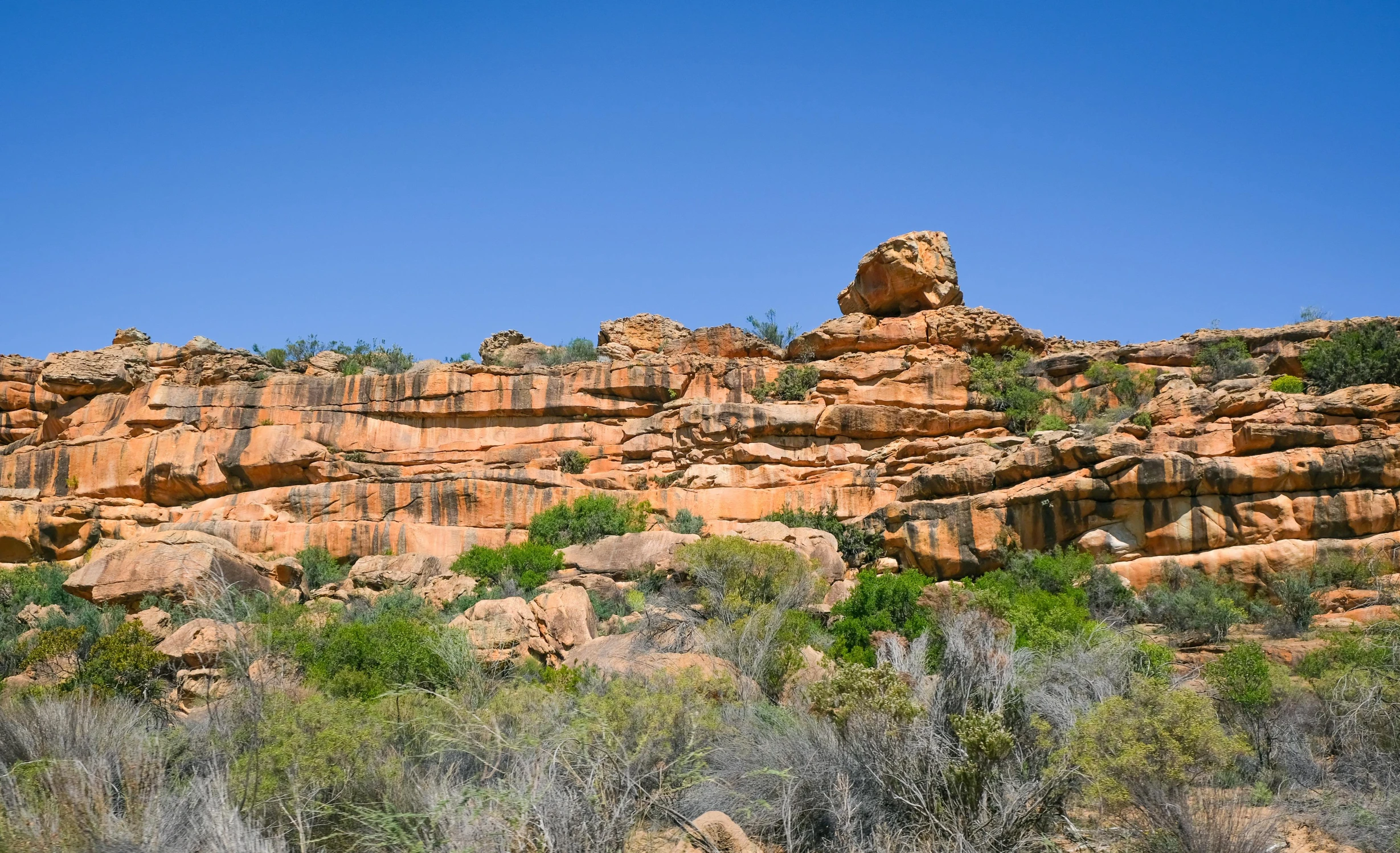 a large rock formation in the middle of a desert, bushveld background, hanging gardens, blue sky, manuka