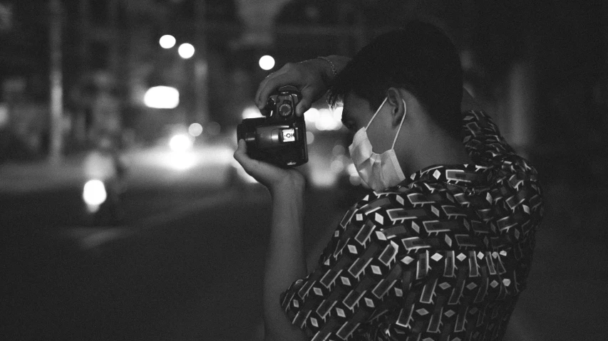 a black and white photo of a woman holding a camera, a black and white photo, inspired by Louis Faurer, unsplash, art photography, the masks come off at night, wearing facemask, night time footage, in style of kar wai wong