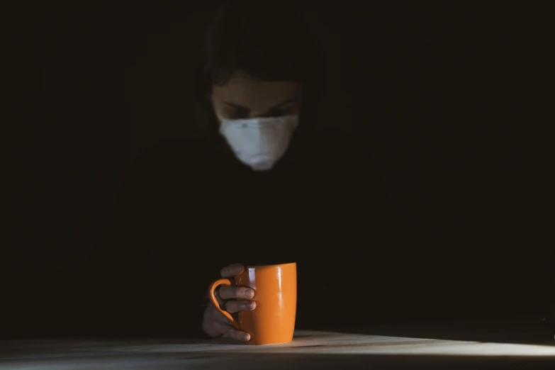 a person sitting at a table with a cup of coffee, partially masked, orange, it's dark, virus