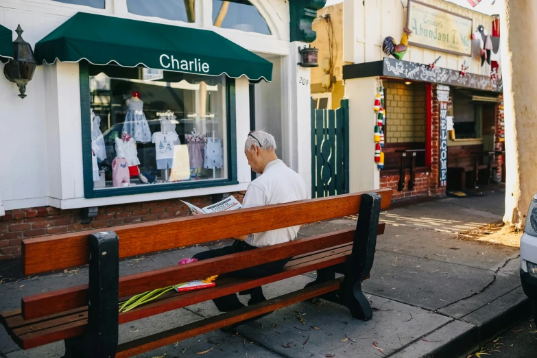 a man sitting on a bench reading a newspaper, a photo, by Charles Martin, pexels contest winner, award winning shopfront design, caulfield, el chavo, sea - green and white clothes