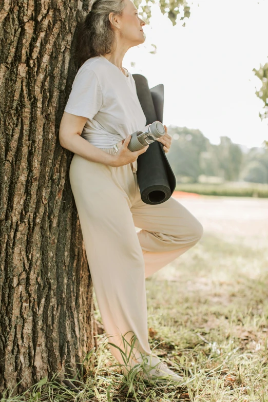 a woman leaning against a tree with a yoga mat, by Carey Morris, pexels contest winner, renaissance, matte black pants, holster, tan, joy ang