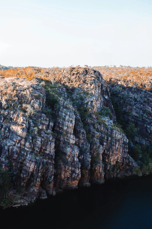 a man standing on top of a cliff next to a body of water, an album cover, unsplash, australian tonalism, highly detailed rock structures, panorama view, high angle vertical, evening sun