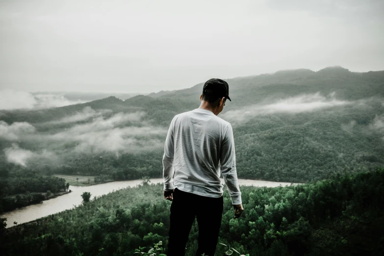 a man standing on top of a lush green hillside, pexels contest winner, overcast gray skies, outlive streetwear collection, man in white t - shirt, looking from behind