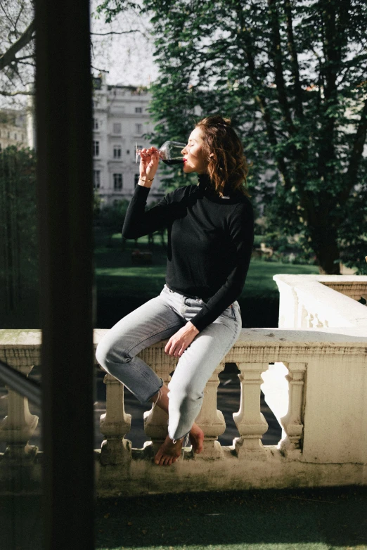 a woman sitting on a balcony drinking from a bottle, by Nina Hamnett, pexels contest winner, happening, black turtle neck shirt, doing an elegant pose over you, full body in frame, promotional image