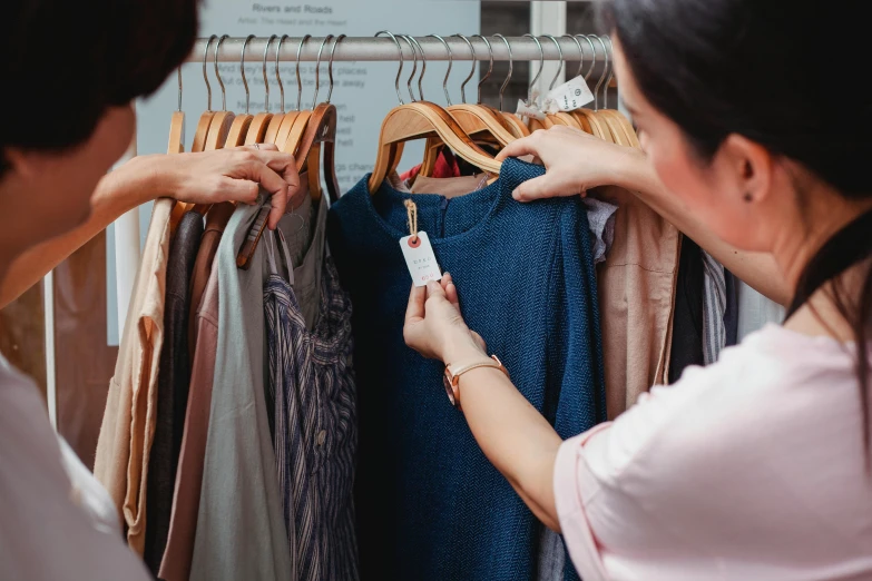 a woman standing in front of a rack of clothes, a photo, trending on pexels, woman holding another woman, at checkout, detaild, highly detaild