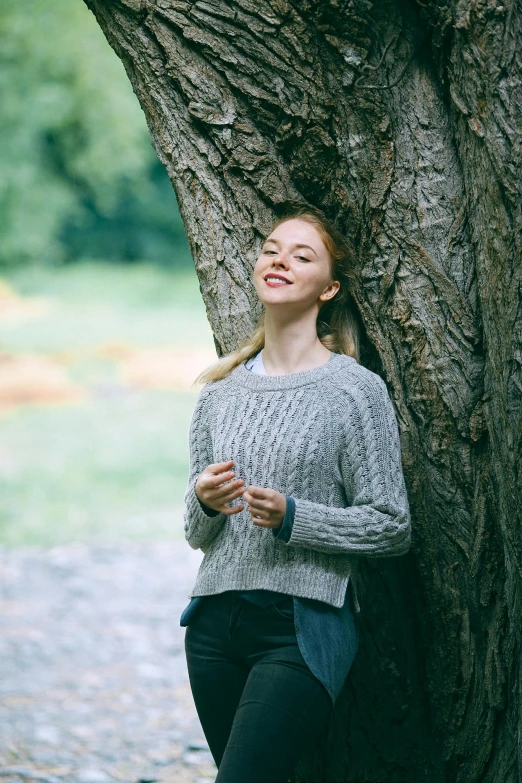 a woman leaning against a tree in a park, inspired by Caroline Lucy Scott, unsplash, renaissance, wearing casual sweater, eleanor tomlinson, celebrating, grey