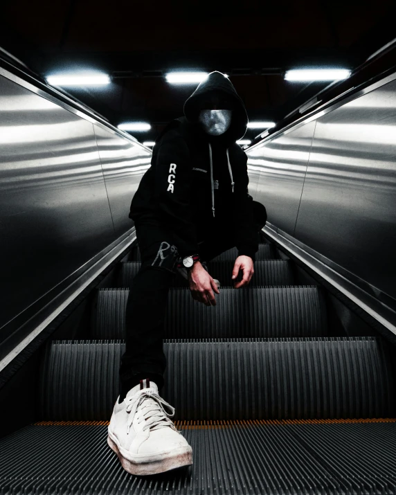 a man in a gas mask sitting on an escalator, an album cover, pexels contest winner, techwear clothes, full body picture, white on black, wearing a black hoodie