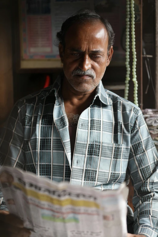 a man sitting at a table reading a newspaper, inspired by Sunil Das, happening, filmstill, portrait image, old charismatic mechanic, promo image