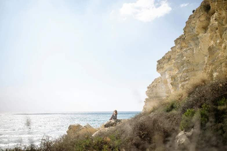 a man sitting on top of a cliff next to the ocean, by Simon Marmion, pexels contest winner, cyprus, photo of a woman, al fresco, without text