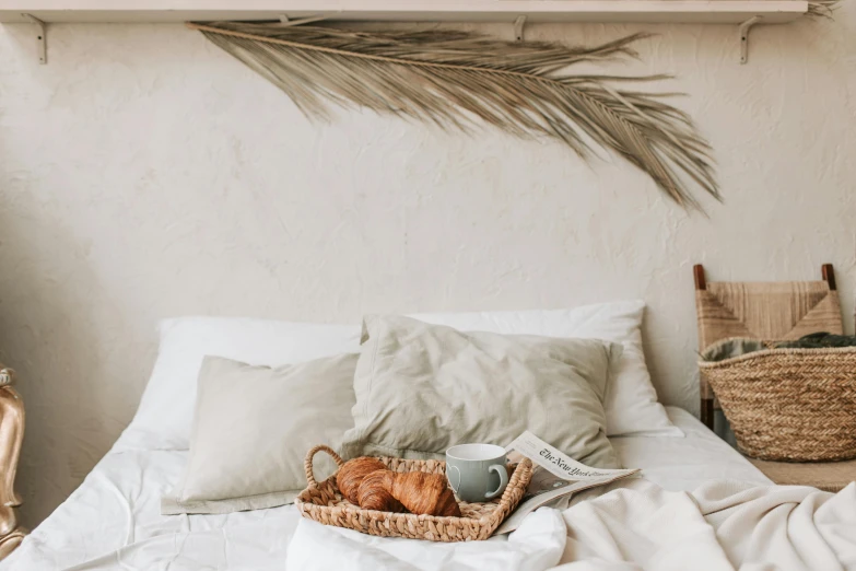 a bed with a basket of croissants on top of it, by Emma Andijewska, pexels contest winner, minimalism, light beige pillows, with a white mug, chest covered with palm leaves, rustic setting