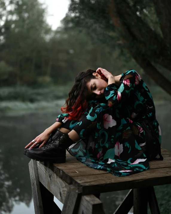 a woman sitting on top of a wooden bench next to a body of water, inspired by Elsa Bleda, pexels contest winner, gothic outfit, floral clothes, stressing out, hugging her knees