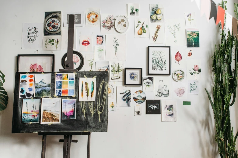 a piece of art hanging on a wall next to a potted plant, a watercolor painting, trending on pexels, inside a cluttered art studio, picture frames, photo of a classroom, set against a white background