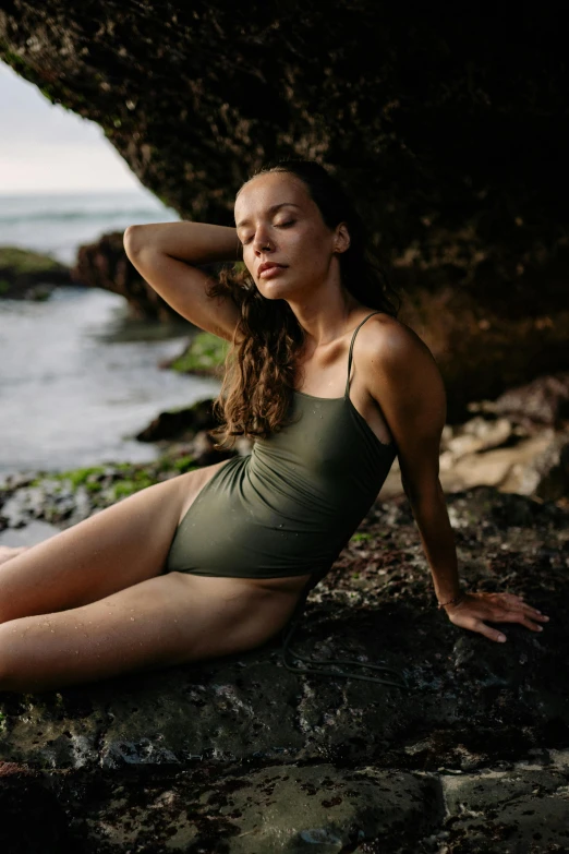 a woman sitting on top of a rock next to the ocean, muted colored bodysuit, olive green, profile image, swimsuit