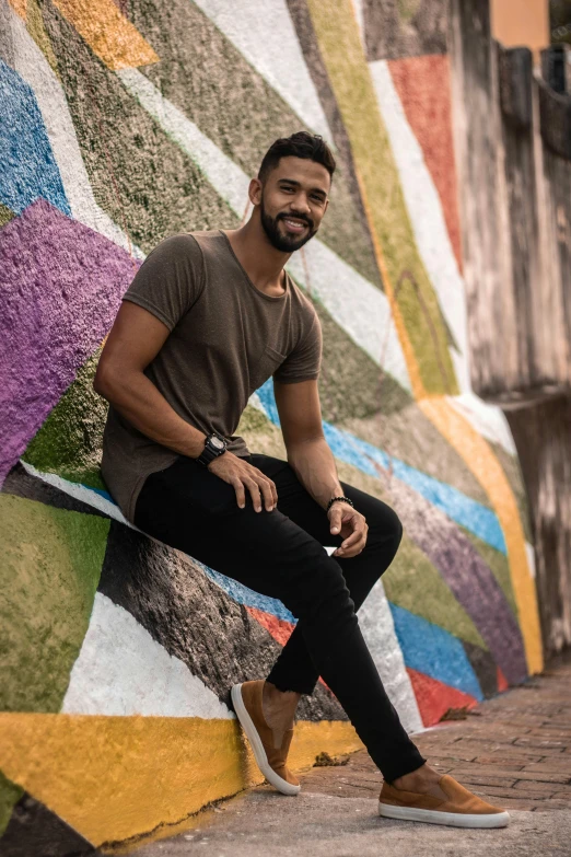 a man sitting on a skateboard in front of a colorful wall, daniel ricciardo, black and brown colors, in sao paulo, smiling man