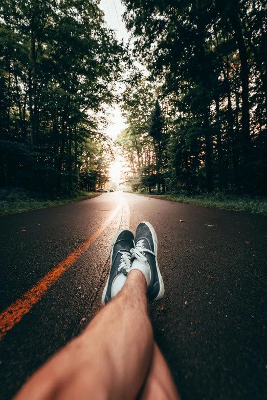 a person riding a skateboard down a road, by Marshall Arisman, unsplash contest winner, realism, lying on the woods path, running shoes, tree-lined path at sunset, selfie photo