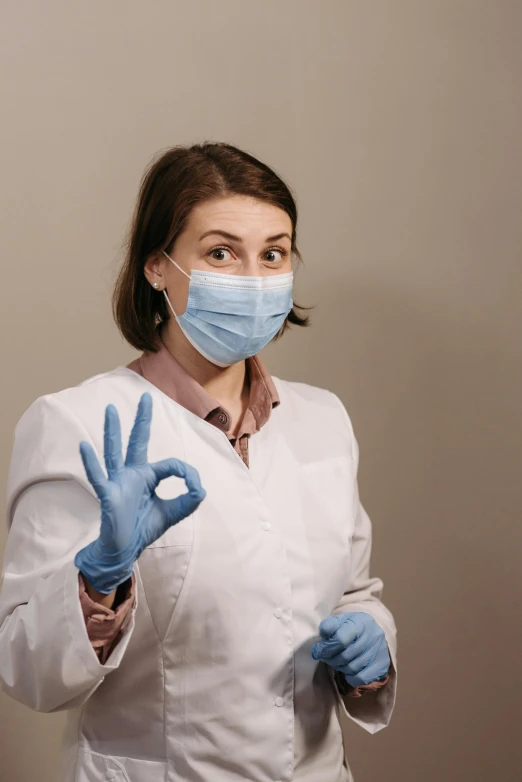 a woman in a white lab coat and blue gloves, a picture, trending on pexels, peace sign, quackery, covid, brown