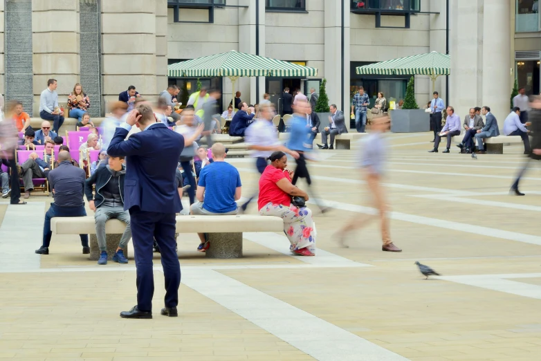 a man in a suit standing in front of a crowd of people, an album cover, inspired by Ruth Orkin, pexels contest winner, coventry city centre, benches, where a large, zoom blur