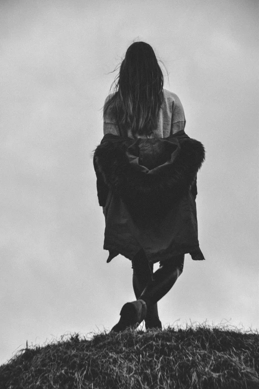 a woman standing on top of a grass covered hill, a black and white photo, by Caro Niederer, unsplash, in a cloak with long hairs, standing on two feet, gloomy and depressed, wearing jacket and skirt