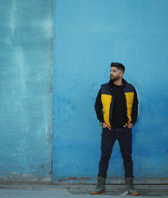 a man standing in front of a blue wall, an album cover, pexels contest winner, yellow navy teal black and gold, reza afshar, wearing a jeans jackets, dark. no text