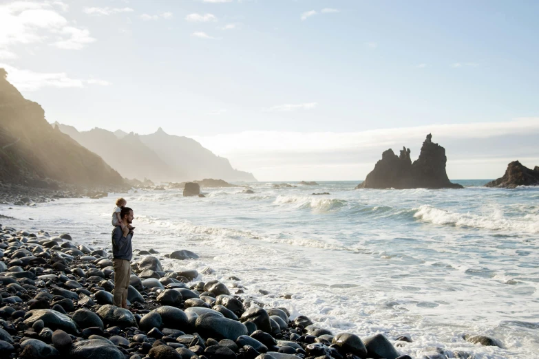 a man standing on a rocky beach next to the ocean, taking a picture, conde nast traveler photo, liam brazier, family