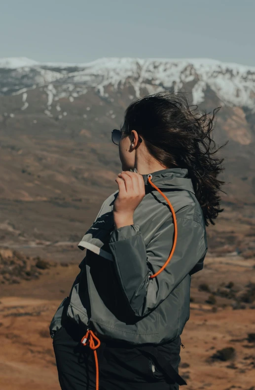 a woman standing on top of a mountain talking on a cell phone, by Andries Stock, trending on unsplash, orange reflective puffy coat, long windy hair style, zippers, low detail