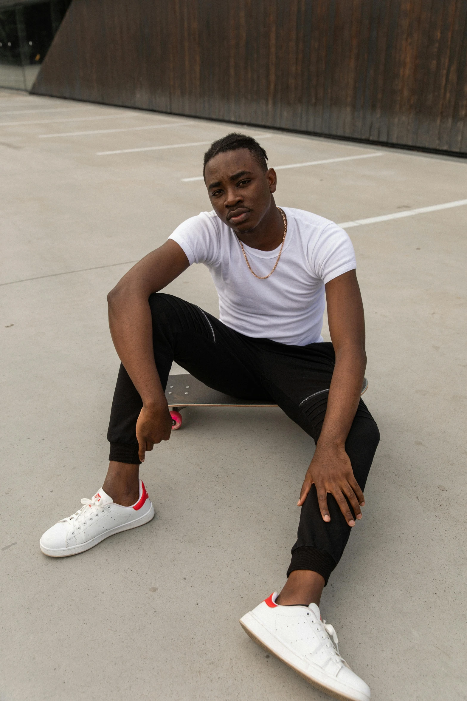 a man sitting on a skateboard in a parking lot, an album cover, by Cosmo Alexander, trending on pexels, dressed in a white t shirt, ( ( dark skin ) ), striking a pose, non-binary