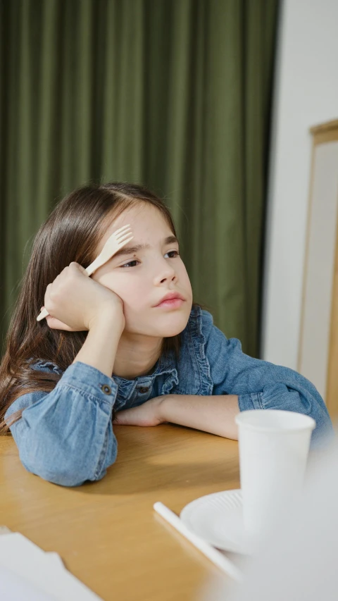 a little girl sitting at a table with a cup of coffee, trending on pexels, realism, bored expression, sitting in the classroom, low quality photo, pondering