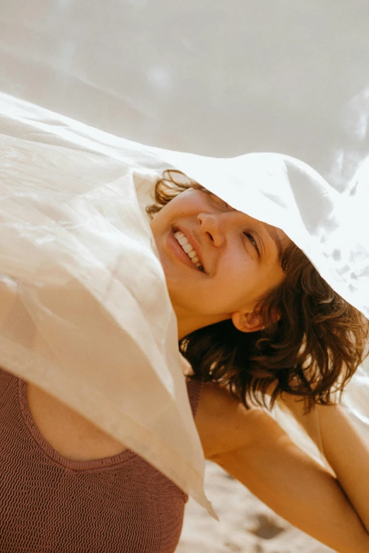 a woman standing on top of a sandy beach, an album cover, by Jessie Algie, trending on unsplash, happening, curled up under the covers, smiling laughing, wearing translucent sheet, closeup of face