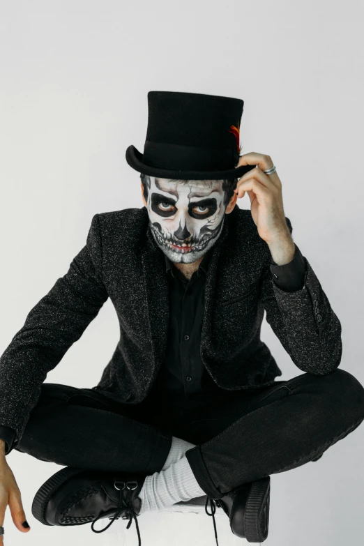 a man wearing a skeleton face paint and a top hat, inspired by James Bolivar Manson, trending on pexels, on a gray background, sitting down casually, kai carpenter, symmetrical head and body