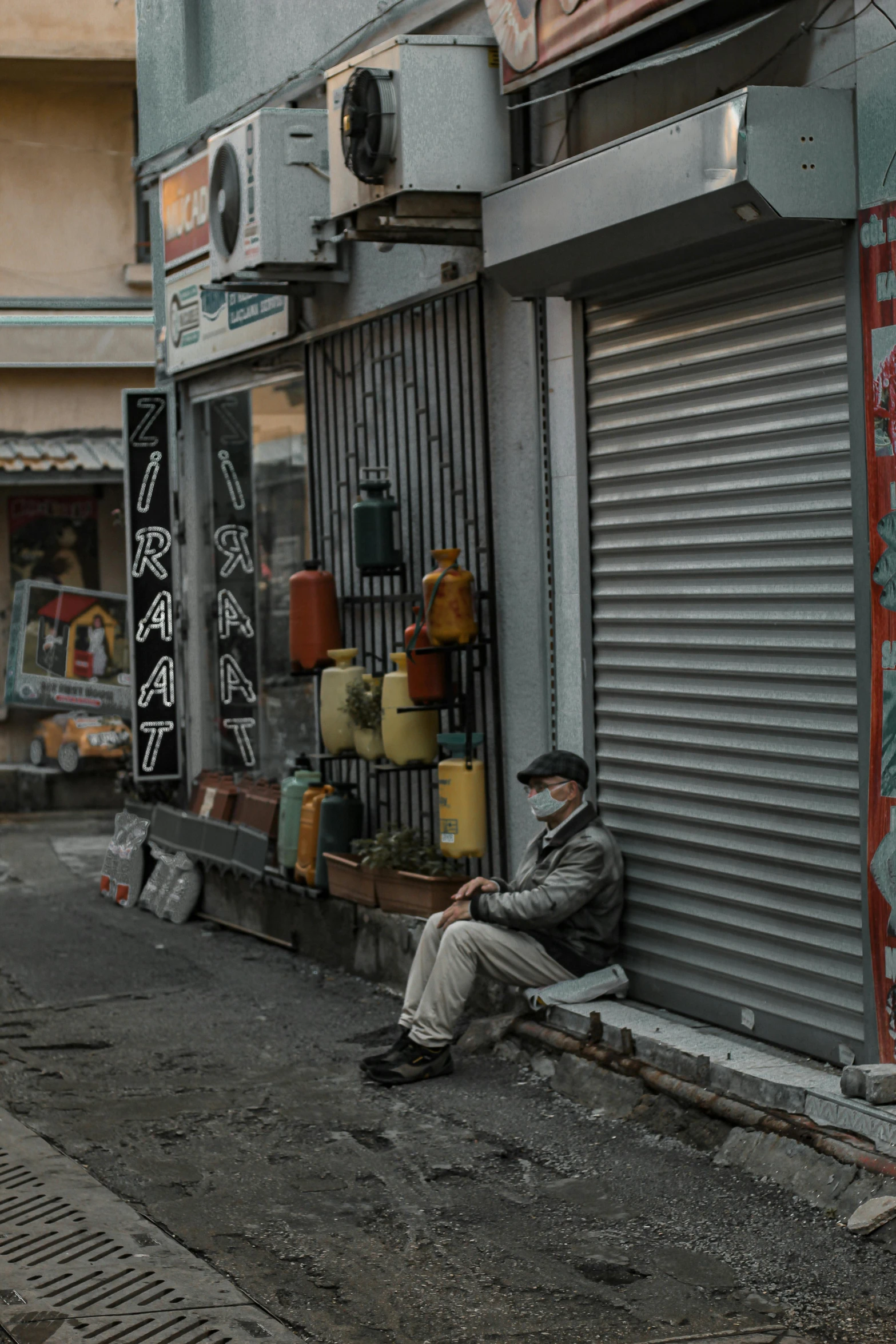 a man sitting on the side of a street next to a building, market in japan, masked person in corner, grey, unsplash photography