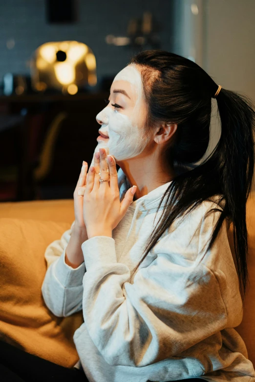 a woman sitting on a couch with a face mask on, by Julia Pishtar, trending on pexels, renaissance, asian beautiful face, doing a prayer, square facial structure, in the bedroom at a sleepover