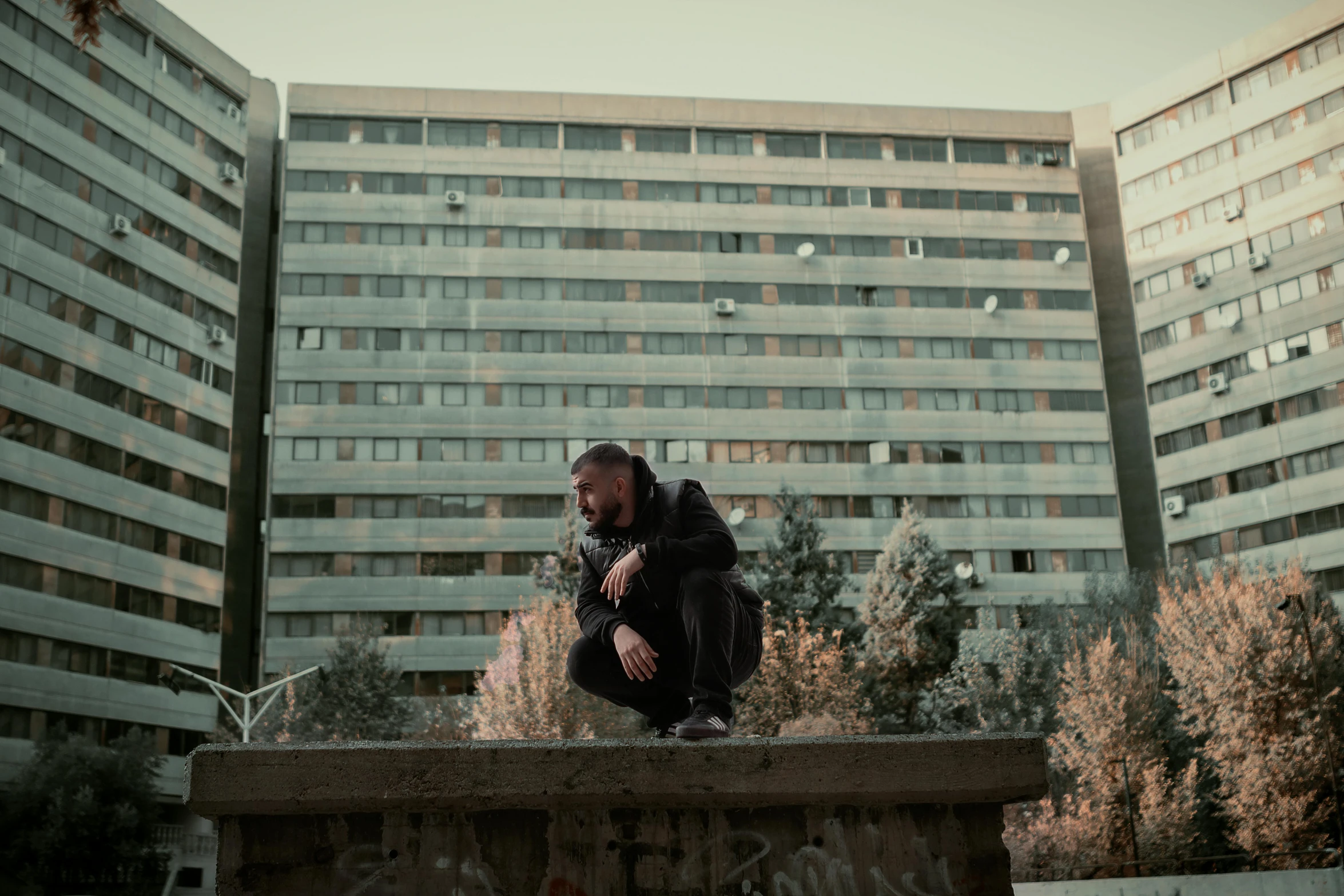 a man sitting on a bench in front of a building, an album cover, pexels contest winner, brutalism, cinematic pose, no people 4k, city buildings on top of trees, crouching