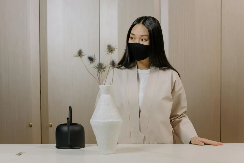 a woman sitting at a table with a vase in front of her, inspired by Kim Tschang Yeul, pexels contest winner, balaclava mask, product design shot, standing elegantly, synthetic materials