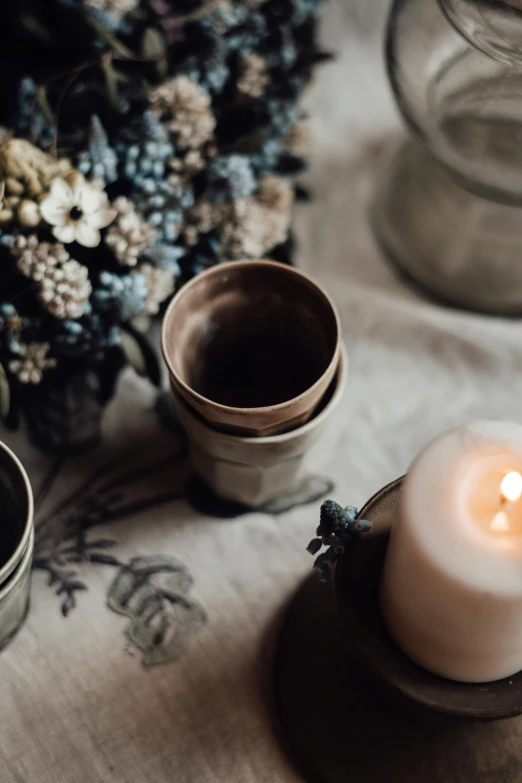 a candle sitting on top of a table next to a vase, a still life, trending on unsplash, wood cups, herbs and flowers, soft light of winter, linen