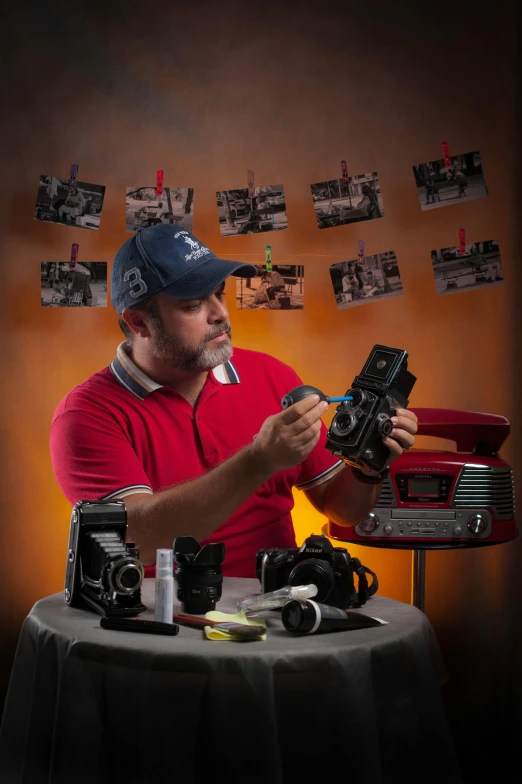 a man sitting at a table holding a camera, an airbrush painting, by Ben Zoeller, with nikon cameras, shot on red camera, in retro colors, toni infante